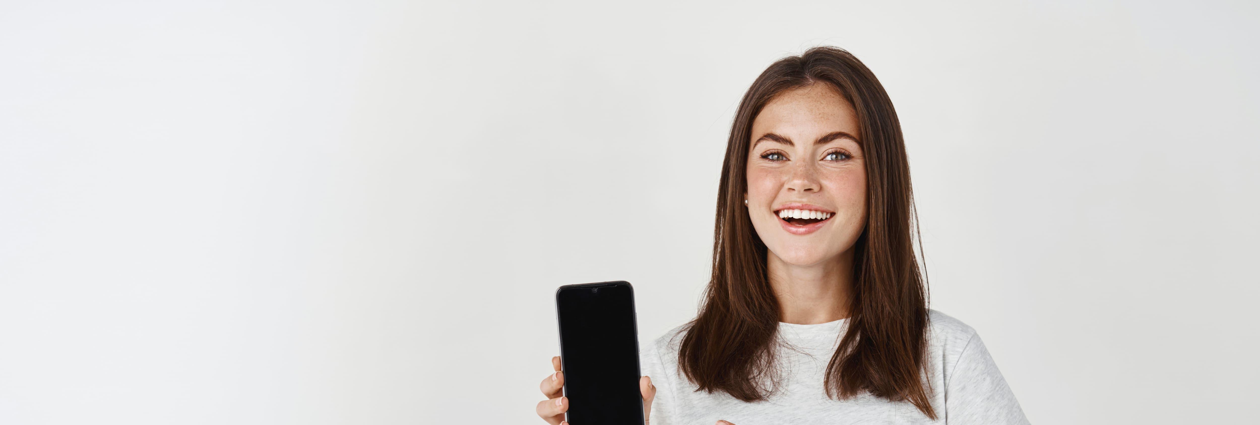 young-brunette-woman-showing-blank-smartphone-screen-pointing-finger-mobile-phone-smiling-satisfied-camera-standing-white-background.jpg